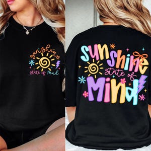 Puede incluir: Camiseta negra con un gráfico colorido que dice "sunshine state of mind". El diseño incluye un sol, un rayo y flores. El mismo diseño está en la parte delantera y trasera de la camiseta.