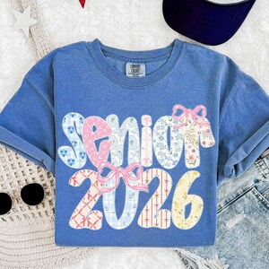 May include: A blue t-shirt with the word "Senior" and the number "2026" in a patchwork design. The word "Senior" is in pink and blue floral patterns, with pink bows. The number "2026" is in various patterns.