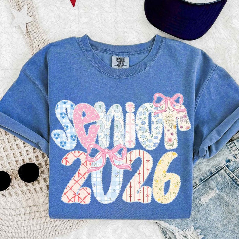May include: A blue t-shirt with the word "Senior" and the number "2026" in a patchwork design. The word "Senior" is in pink and blue floral patterns, with pink bows. The number "2026" is in various patterns.