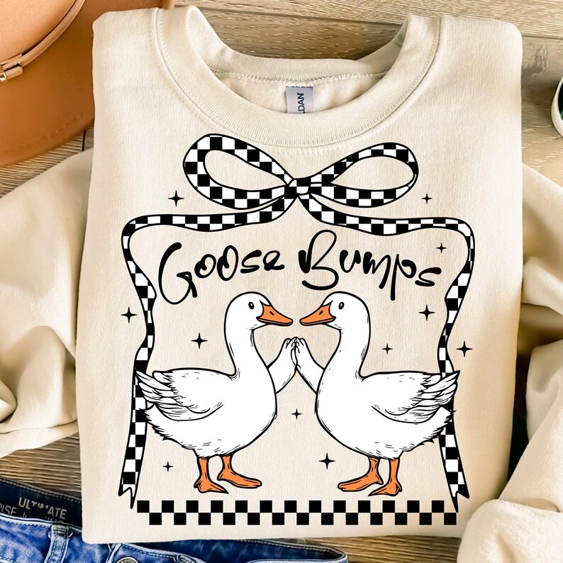 May include: A cream-coloured sweatshirt with a black and white chequered bow and the text "Goosebumps" in black. Two white geese are facing each other with their wings outstretched.