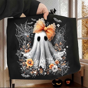 May include: Black Halloween-themed decorative pillow featuring a white ghost with an orange bow, surrounded by spiderwebs, flowers, and bats. The ghost has black eyes and is wearing a lace collar. The pillow is held up by a person.