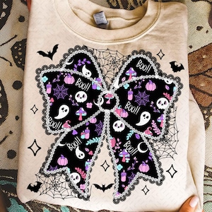 May include: Beige sweatshirt with a large black bow design. The bow is decorated with Halloween-themed illustrations, including ghosts, skulls, pumpkins, and the word "Boo!!". The bow is outlined with a lace-like trim and surrounded by spiderwebs and bats.