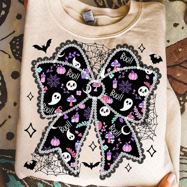 May include: Beige sweatshirt with a large black bow design. The bow is decorated with Halloween-themed illustrations, including ghosts, skulls, pumpkins, and the word "Boo!!". The bow is outlined with a lace-like trim and surrounded by spiderwebs and bats.
