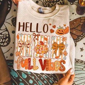 Puede incluir: Sudadera beige con un diseño de temática otoñal. El diseño incluye las palabras "HELLO FALL VIBES" en marrón y naranja, junto con calabazas, lazos y hojas. La sudadera está doblada y sostenida por manos.