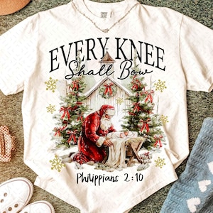 May include: White t-shirt featuring a Christmas scene with Santa kneeling before a manger. The shirt has the text "EVERY KNEE SHALL BOW" and "Philippians 2:10". The design includes Christmas trees, snowflakes, and red bows.