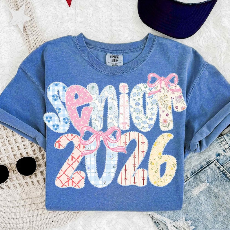 May include: Blue t-shirt with the word "Senior 2026" in patchwork letters and numbers. The letters and numbers are decorated with floral and plaid patterns, and pink bows. The shirt is a Comfort Colors brand.