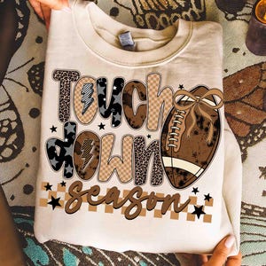May include: Cream-colored sweatshirt with the words "Touch Down Season" in a mix of fonts and patterns, including leopard print and checkered designs. A brown football with a bow is featured on the right side, with black star accents.