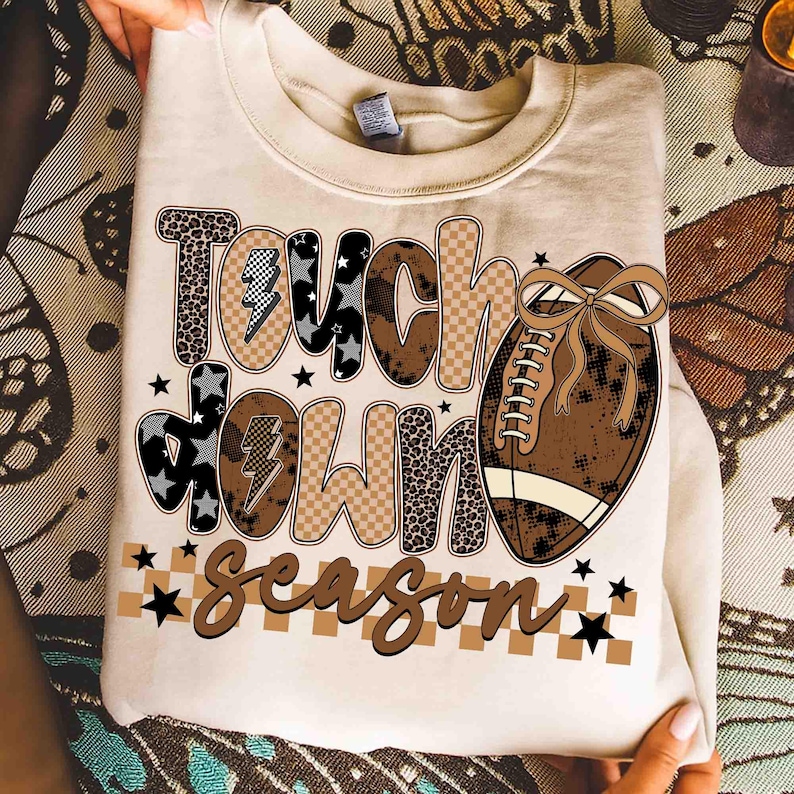 May include: Cream-colored sweatshirt with the words "Touch Down Season" in a mix of fonts and patterns, including leopard print and checkered designs. A brown football with a bow is featured on the right side, with black star accents.