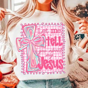 May include: White sweatshirt featuring a pink cross design with a bow and the text "Let me tell you about my Jesus." The cross has blue and pink swirls. The background is a pink and white gingham pattern.
