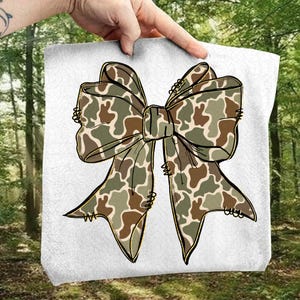 May include: A decorative bow with a camouflage pattern in shades of green, brown, and tan. The bow is centered on a white background and held by a hand. The bow's design features detailed edges and a classic bow shape.