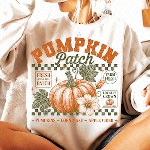 Può includere: Una felpa beige a girocollo con una stampa grafica che riporta il testo PUMPKIN PATCH in arancione, un motivo a scacchi, una grande zucca arancione, foglie verdi, un fiore e riquadri con le scritte FRESH FROM THE PATCH, FARM FRESH, LOCALLY GROWN.