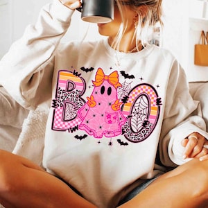 May include: A cream-colored sweatshirt featuring a pink ghost with an orange bow, peace signs, and a flower pocket. The word "BOO" is in a mix of leopard print, orange, and pink, with spiderwebs and bats. A fun Halloween-themed design.