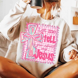 May include: Cream-colored sweatshirt featuring a pink and blue cross design with the text "Let me tell you about my Jesus." The cross has a bow and is set against a pink and white checkered background.