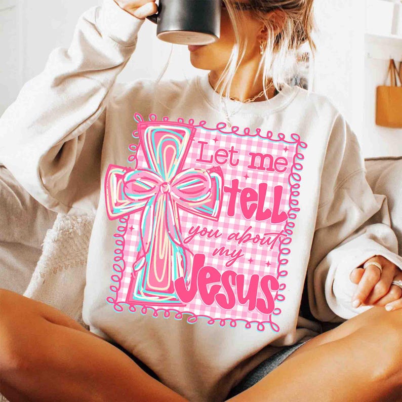 May include: Cream-colored sweatshirt featuring a pink and blue cross design with the text "Let me tell you about my Jesus." The cross has a bow and is set against a pink and white checkered background.