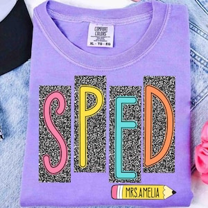 May include: A lavender Comfort Colors t-shirt with the word "SPED" in colorful block letters. The letters are set against a black and white speckled background. A pencil graphic with the text "MRS. AMELIA" is below the letters.