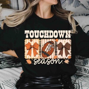 May include: Black long-sleeve shirt with a graphic that says "TOUCHDOWN season." The graphic includes a football, bows, and fall leaves in shades of orange, brown, and white. The shirt is made of a soft material.