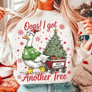 May include: White sweatshirt with a whimsical Christmas design. It features a goose wearing a Santa hat, green sweater, and yellow boots, pulling a cart with a decorated Christmas tree. Text includes "Oops! I got" and "Another Tree".