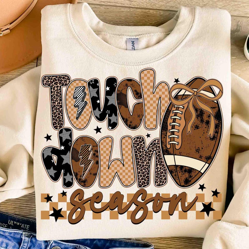 May include: Cream-colored sweatshirt with the words "Touch Down Season" in a mix of leopard print, checkered, and star patterns. A brown football with a bow is on the right side. Black stars are scattered around the text.