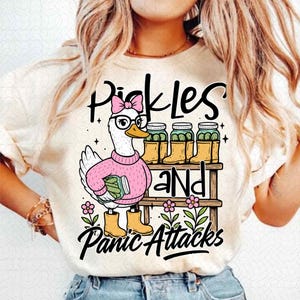 May include: Cream-colored t-shirt featuring a cartoon duck wearing glasses, a pink sweater, and yellow boots. The duck is holding a jar of pickles and standing near a shelf with jars of pickles. Text on the shirt reads "Pickles and Panic Attacks."