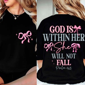 Op de afbeelding: Zwart T-shirt met een roze strikpatroon en de tekst "GOD IS WITHIN HER She WILL NOT FALL Psalm 46:5". Het shirt heeft een ronde hals en korte mouwen. De strik is ook op de voorkant.
