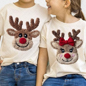 May include: Two cream-colored t-shirts with reindeer faces embroidered on the front. The reindeer have brown antlers, a red nose, and a smiling mouth. One reindeer has a red bow. The shirts are paired with blue jeans.