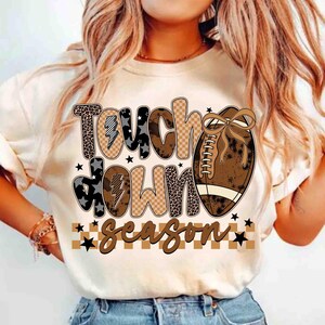 May include: A cream-colored t-shirt featuring the words "Touch Down Season" in a mix of patterned fonts and a football graphic. The design includes leopard print, stars, and a brown and tan color scheme, perfect for football fans.