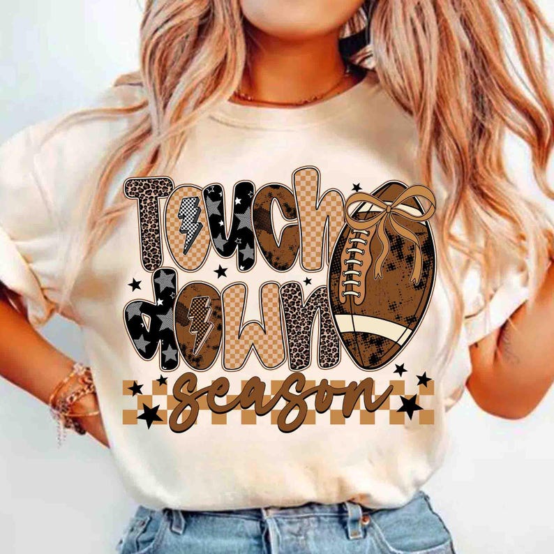 May include: A cream-colored t-shirt featuring the words "Touch Down Season" in a mix of patterned fonts and a football graphic. The design includes leopard print, stars, and a brown and tan color scheme, perfect for football fans.