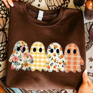May include: A brown sweatshirt featuring four ghost designs with black eyes. The ghosts have different patterns: floral, yellow gingham, floral with orange accents, and orange plaid. The sweatshirt is held by hands.