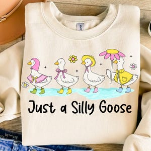 May include: A beige sweatshirt with a graphic of four geese wearing hats and carrying umbrellas. The geese are wearing pink, yellow, and blue accessories. The text "Just a Silly Goose" is printed below the geese.