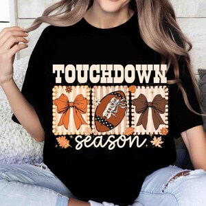 May include: Black t-shirt with a football-themed graphic. The design includes the word "TOUCHDOWN" above three rectangular panels. The panels feature a bow, a football, and another bow, with the word "season" below.