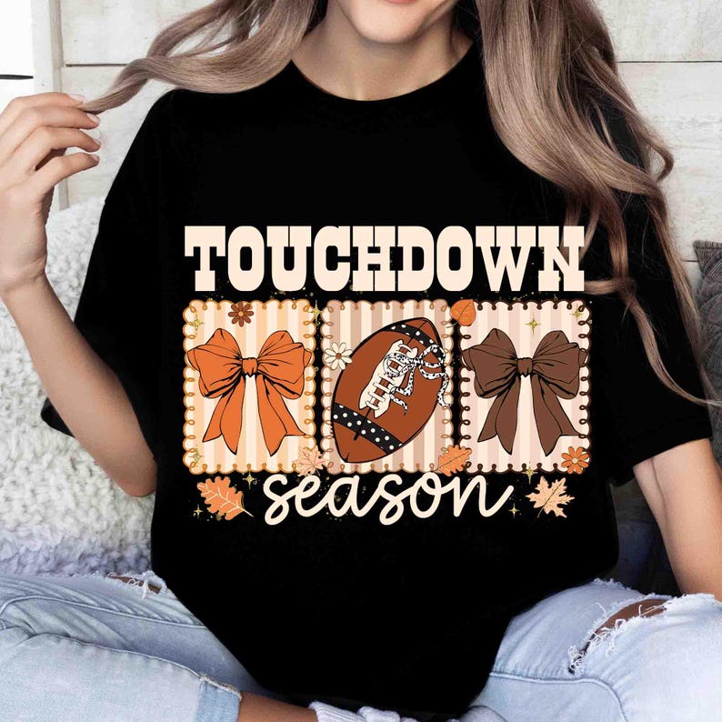 May include: Black t-shirt with a football-themed graphic. The design includes the word "TOUCHDOWN" above three rectangular panels. The panels feature a bow, a football, and another bow, with the word "season" below.