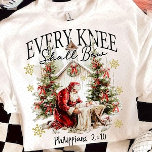 May include: White t-shirt featuring a Christmas scene with Santa kneeling before baby Jesus in a manger. The text "EVERY KNEE SHALL BOW" and "Philippians 2:10" are printed above and below the image, with Christmas trees and snowflakes.