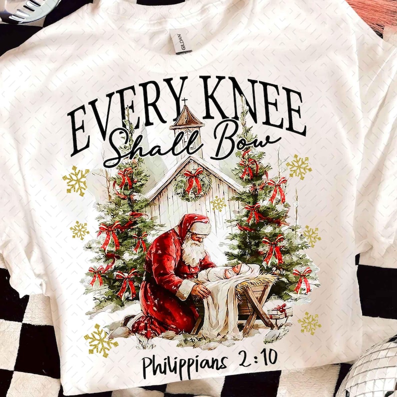 May include: White t-shirt featuring a Christmas scene with Santa kneeling before baby Jesus in a manger. The text "EVERY KNEE SHALL BOW" and "Philippians 2:10" are printed above and below the image, with Christmas trees and snowflakes.