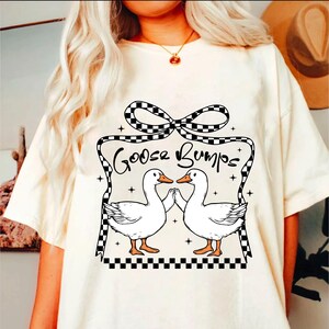 May include: White t-shirt featuring a black and white checked bow and the text "Goose Bumps". Two white geese with orange beaks are facing each other with their wings outstretched.