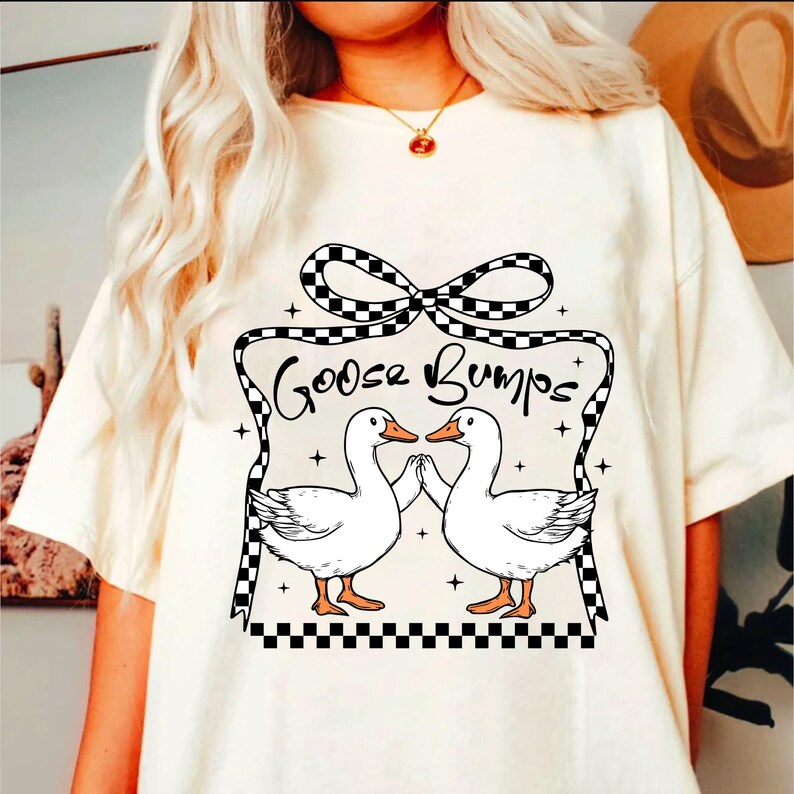 May include: White t-shirt featuring a black and white checked bow and the text "Goose Bumps". Two white geese with orange beaks are facing each other with their wings outstretched.