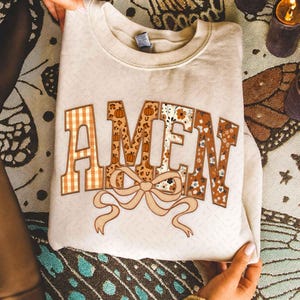 Puede incluir: Una sudadera de color crema con la palabra "AMEN" en una fuente decorativa. Las letras presentan varios patrones, incluyendo cuadros, estampado de leopardo, calabazas y diseños florales. Debajo de la palabra hay un lazo beige.