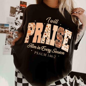 May include: Black t-shirt with the words "I will PRAISE Him in Every Season" in a fall-themed design. The word "PRAISE" features a mix of floral, plaid, and leaf patterns. Psalm 146:2 is printed below the main text.