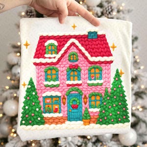 May include: A pink and red Christmas house design with a knitted texture. The house features a red roof, blue door, and green Christmas trees on either side. The windows have yellow frames and the house is decorated with festive details.