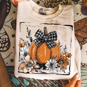 May include: A cream-colored sweatshirt featuring a large orange pumpkin with a black and white plaid bow. The pumpkin is surrounded by white and orange flowers and greenery, all within a black rectangular frame. A fall-themed design.
