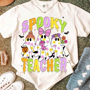 May include: Cream-coloured t-shirt with the words "Spooky Teacher" in a colourful, dripping font. The graphic features three ghost characters decorated with flowers, bows, and accessories. Bats and stars complete the design.