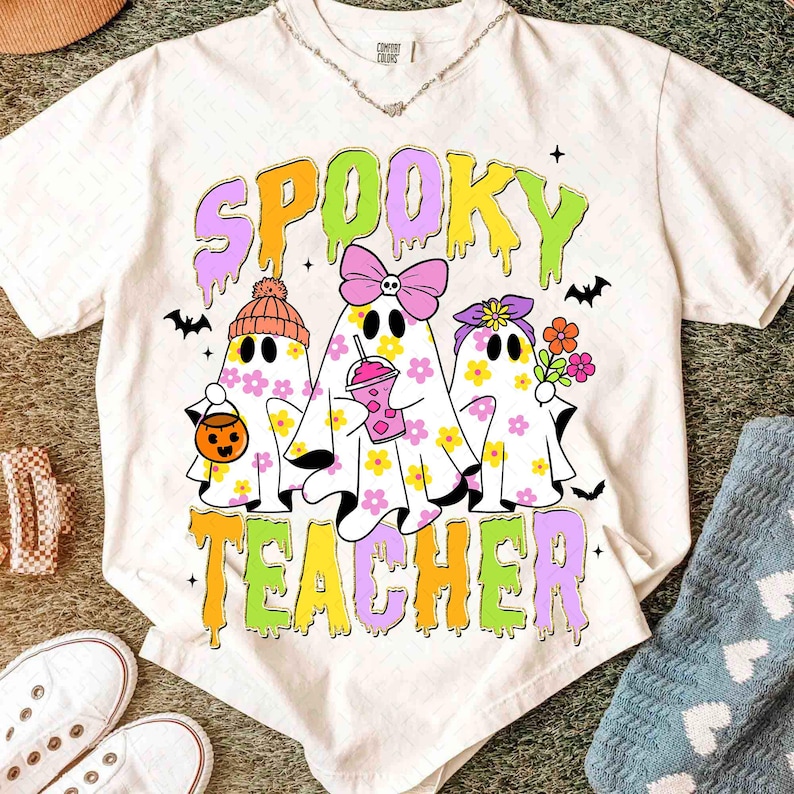 May include: Cream-coloured t-shirt with the words "Spooky Teacher" in a colourful, dripping font. The graphic features three ghost characters decorated with flowers, bows, and accessories. Bats and stars complete the design.