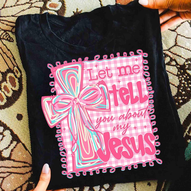 May include: Black t-shirt featuring a pink and blue cross design with the text "Let me tell you about my Jesus." The cross has a bow and is set against a pink and white gingham background.