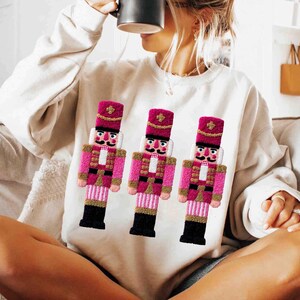 Könnte beinhalten: Cremefarbenes Sweatshirt mit drei gestickten rosa-goldenen Nussknackerfiguren. Die Nussknacker haben schwarze Stiefel, weiße Gesichter und tragen rosa Hüte. Das Sweatshirt ist ein lässiges, festliches Kleidungsstück.