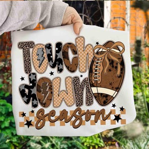 May include: White tote bag featuring the words "Touch Down Season" in a mix of fonts and patterns, including leopard print, stars, and a football graphic. The football is brown with a bow. Black stars and a brown and white checkered pattern complete the design.