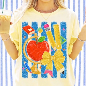 Peut inclure: Un t-shirt jaune pâle avec une impression graphique représentant une pomme rouge, un globe doré, un crayon jaune, une règle jaune, une petite gomme verte et un livre rouge. Le motif est sur un fond à rayures verticales bleues et blanches.