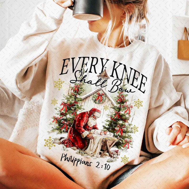 May include: Cream-colored sweatshirt featuring a Christmas-themed graphic. The design includes text that reads "EVERY KNEE Shall Bow" above an image of Santa Claus kneeling before a manger. The text "Philippians 2:10" is printed below the image.
