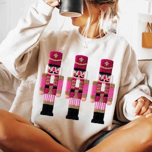 May include: Cream-colored sweatshirt featuring three embroidered pink and gold nutcracker figures. The nutcrackers have black boots, white faces, and are wearing pink hats. The sweatshirt is a casual, festive garment.