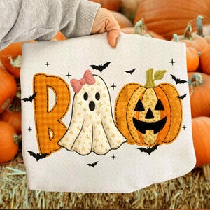 May include: A Halloween-themed decorative pillow featuring the word "BOO" with a pumpkin and ghost design. The "B" is orange plaid, the ghost has a pink bow, and the pumpkin has a carved face. Black bats and plus signs surround the design.