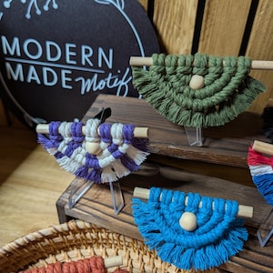 May include: A collection of small, semi-circular macrame wall hangings, each featuring a wooden dowel, a central light-coloured wooden bead, and fringed edges. The pieces are displayed in various solid colours including olive green, blue, and rust.