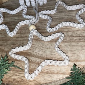 May include: Close-up of three white macrame star ornaments with wooden bead accents. The stars are arranged on a wooden surface, with greenery in the corners. These handmade stars are perfect for holiday decor.